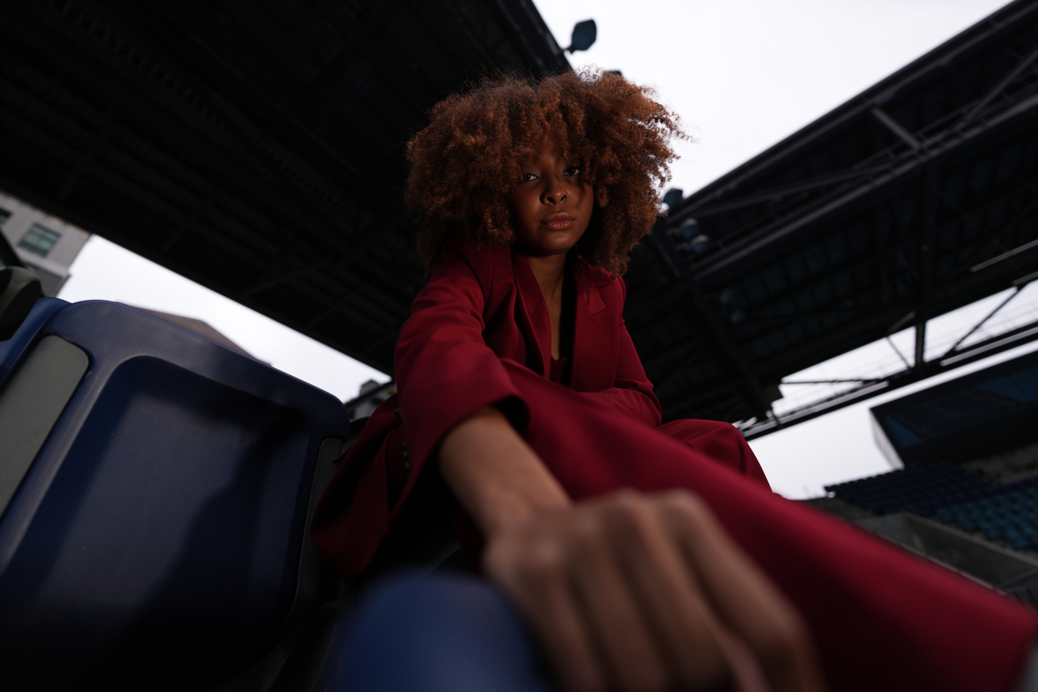 A photograph taken from a low position, featuring a woman in a red suit seated in a blue chair. Her face is positioned small in the centre, with her right hand placed in the foreground, creating a dynamic composition. Click to open modal