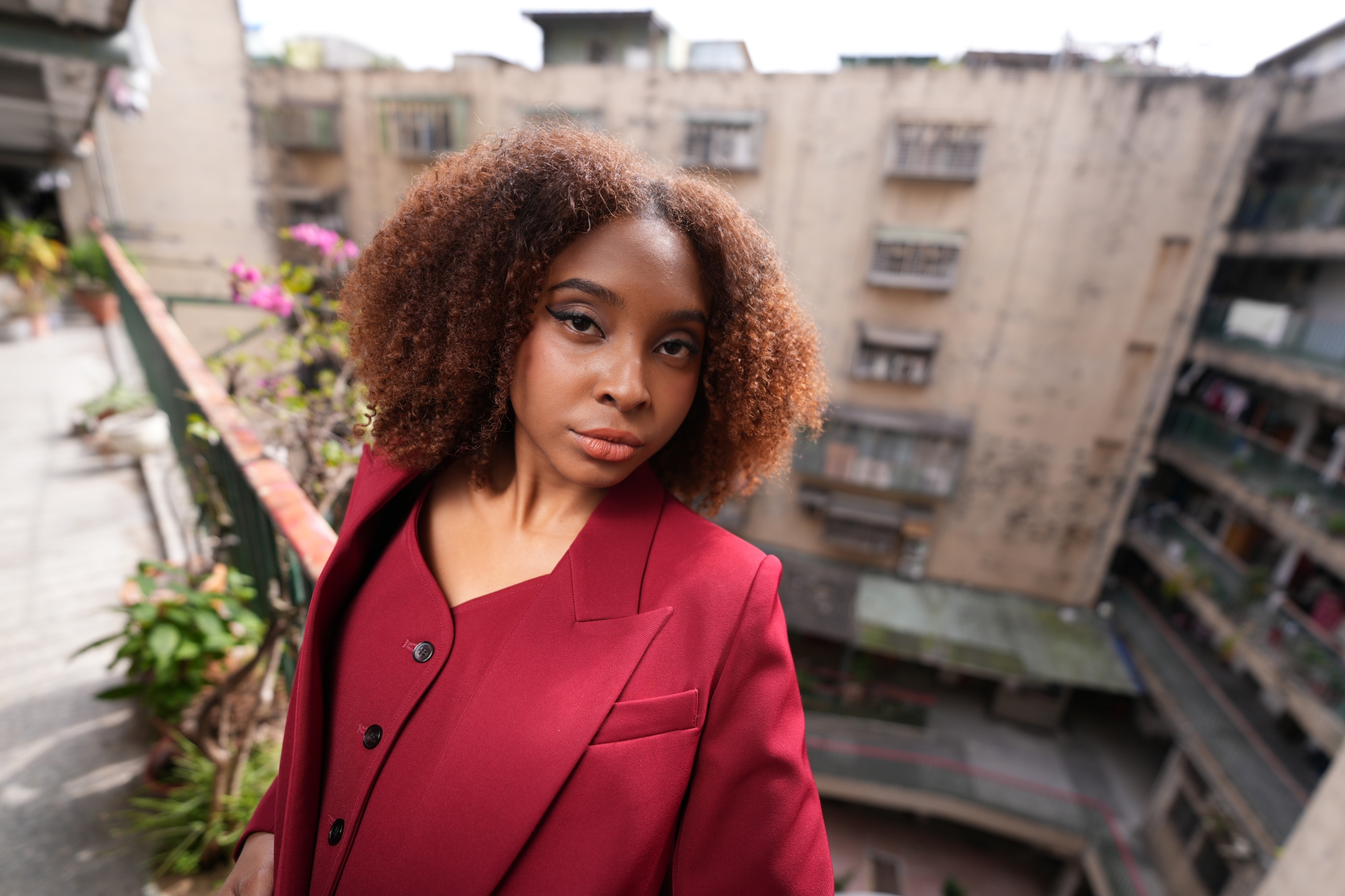 A high-resolution photograph taken from a high position, featuring a woman in a red suit shown from the waist up. Sunlight falls on her right cheek, while the background displays an old, spacious Taiwanese apartment and a deep pink nose, beautifully Bokeh-ed, with the woman leaning against the red railing of the apartment. Click to open modal