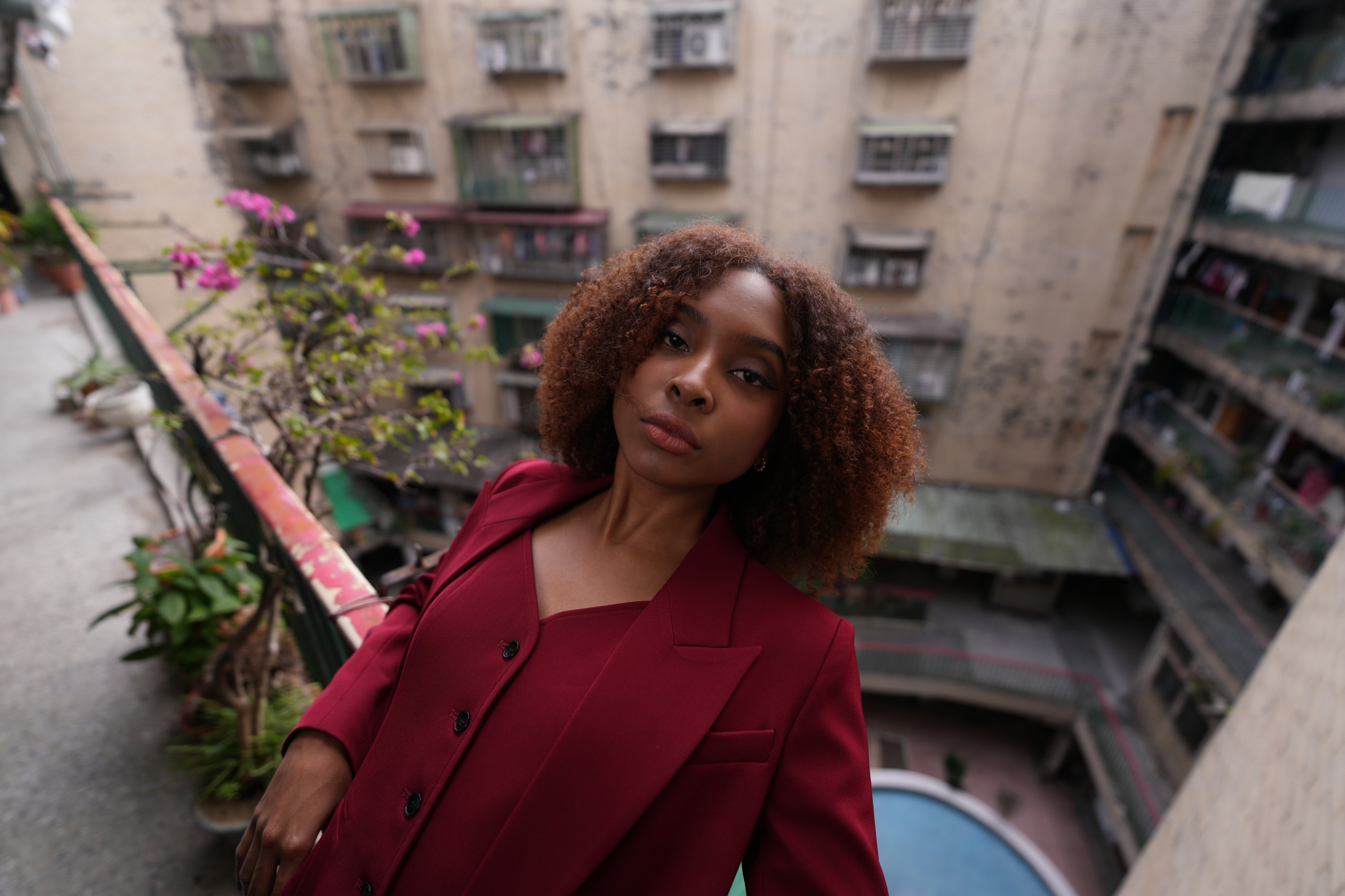 A woman in a red suit is captured in high resolution from a high angle, showing her from the waist up. In the background, an old, spacious Taiwanese apartment and deep pink flowers are beautifully defocused, with the woman leaning against the red railing of the apartment. Click to open modal