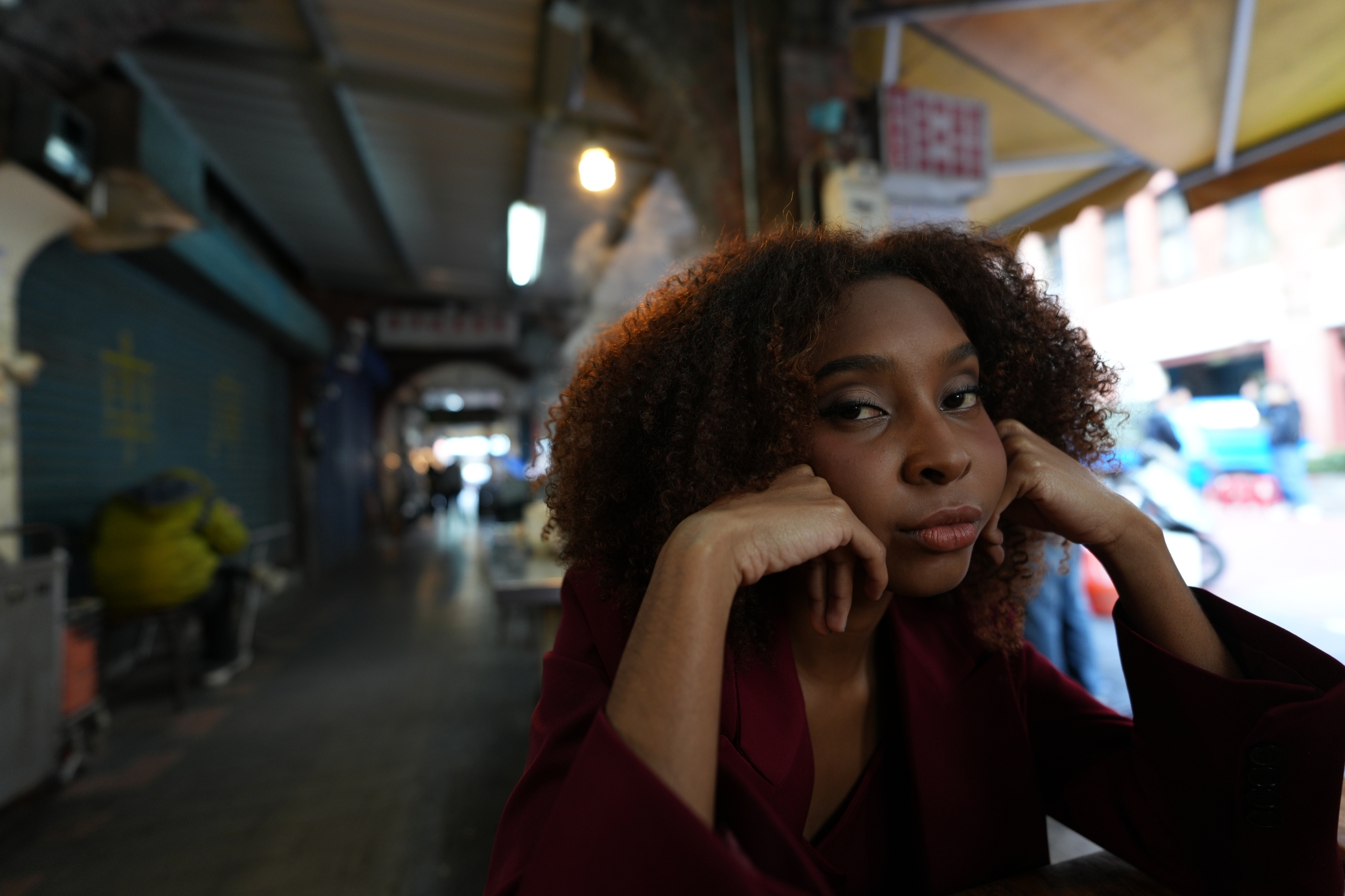 A photograph of a cool woman in a red suit resting her chin on her hand, gazing towards the lens at a table in a corridor featuring typical Asian street stalls. The background corridor is beautifully defocused, creating a sense of depth. Click to open modal