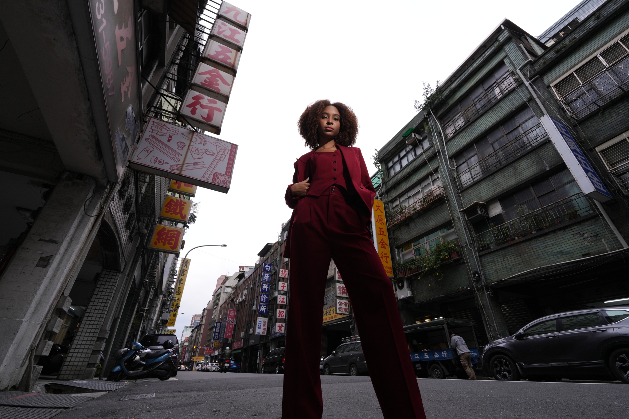 A high-resolution photograph taken from a low position, capturing the full body of a cool woman in a red suit on a street adorned with unique Chinese characters signs on either side. Click to open modal