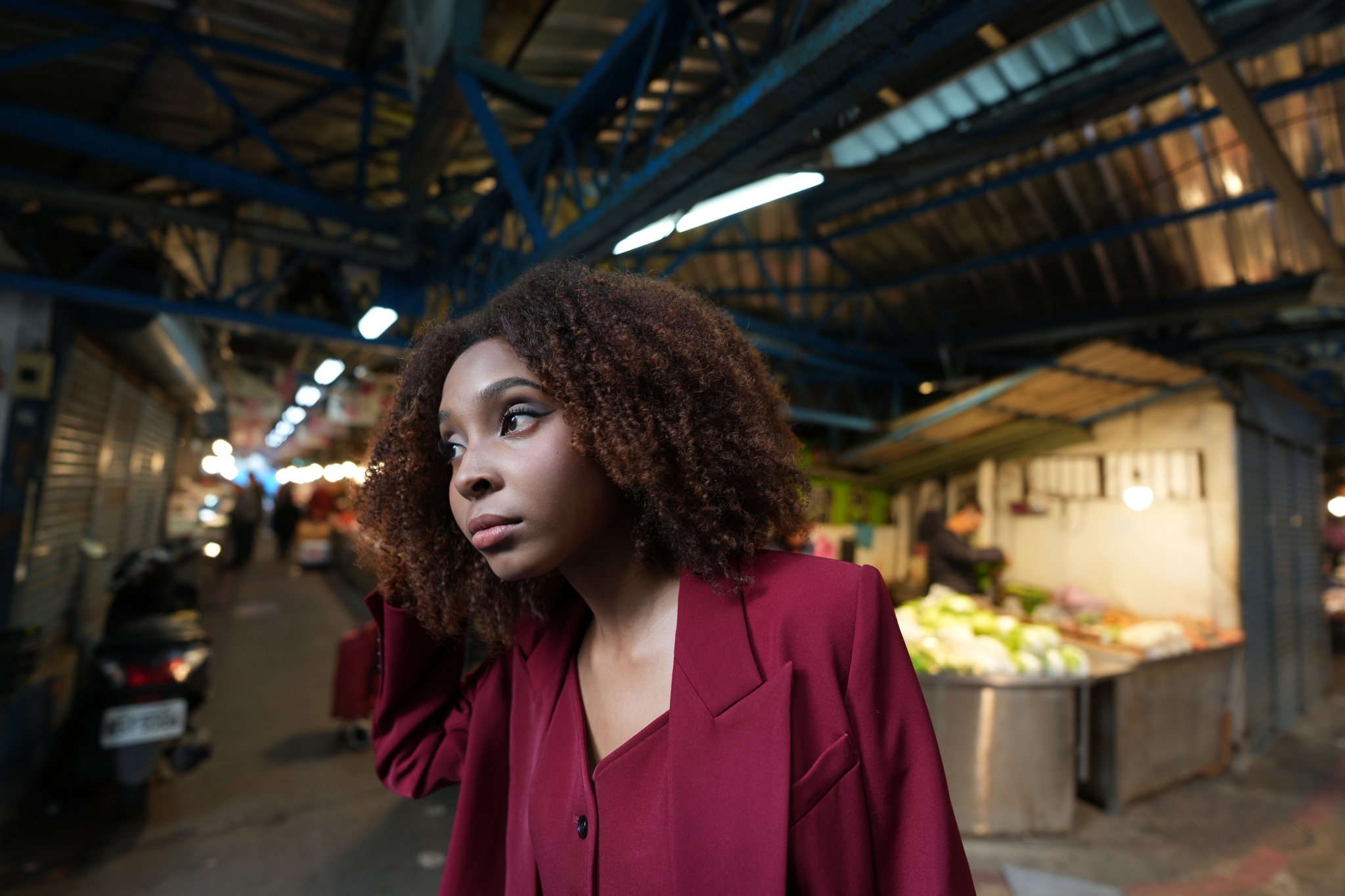 A photograph featuring a woman in a red suit, captured in a bust-up shot at the centre, with her right hand placed behind her head. Her face is in high resolution, while the market scene on either side is beautifully Bokeh-ed, creating a sense of depth. Click to open modal