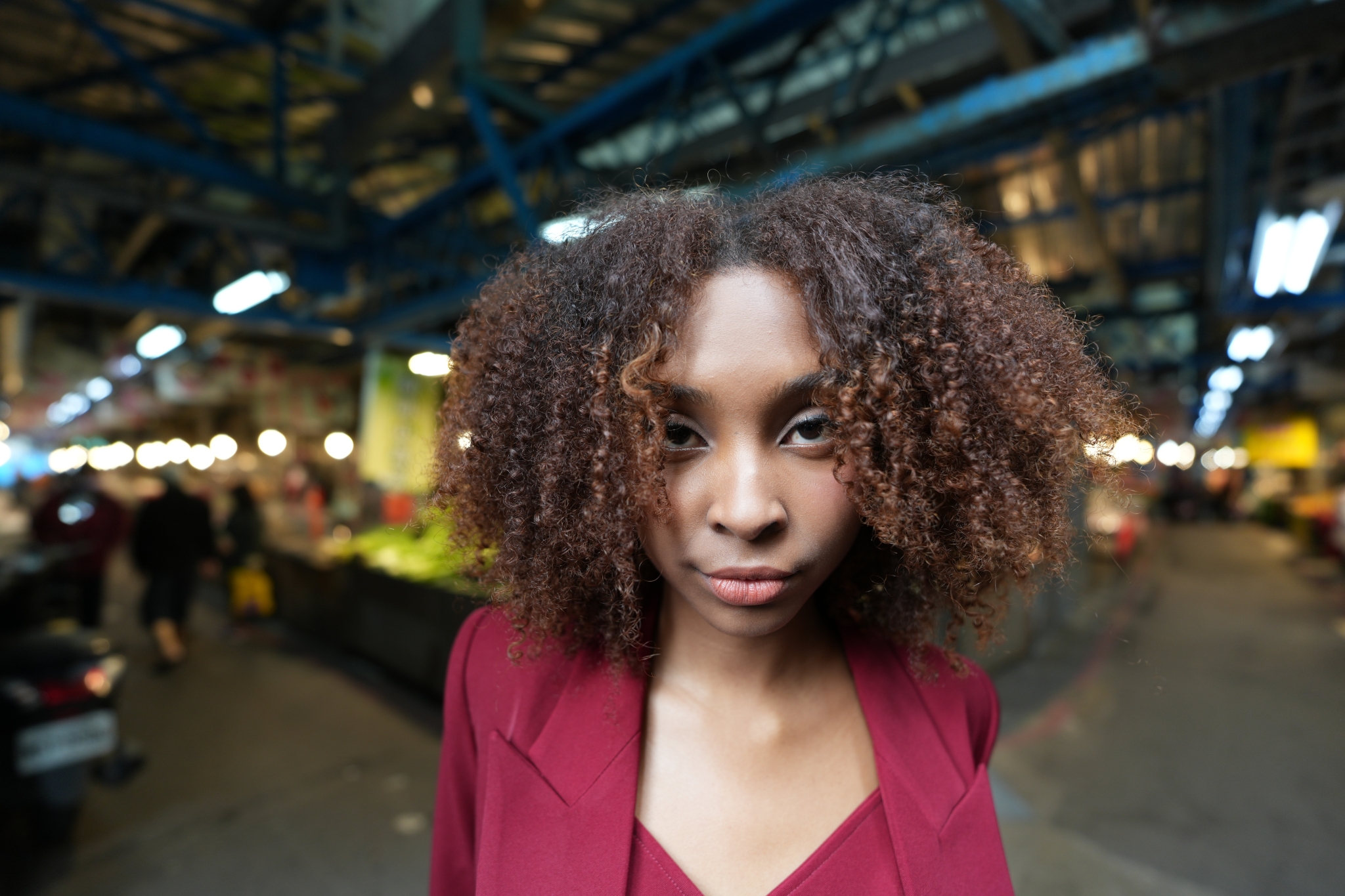 A photograph featuring a woman in a red suit, captured in a bust-up shot at the centre. Her face is in high resolution, while the market scene on either side is beautifully defocused, creating a sense of depth. Click to open modal