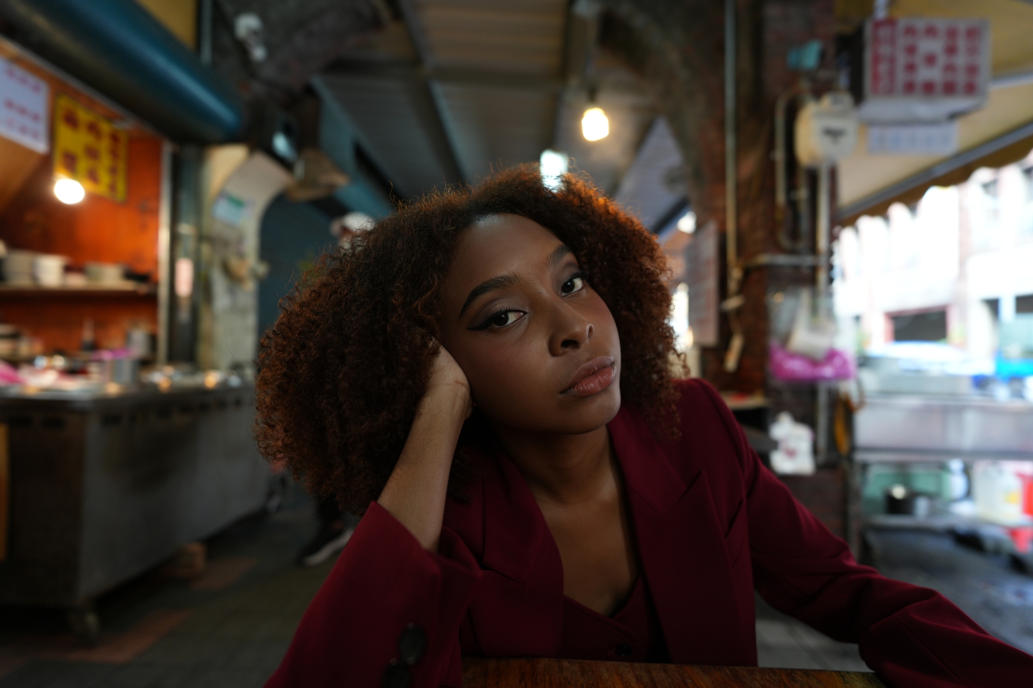 A photograph of a woman in a red suit sitting at a table in a corridor featuring typical Asian street stalls, resting her cheek on her hand and gazing provocatively at the lens. The background corridor is beautifully Bokeh-ed, while the woman is captured in high resolution. Click to open modal