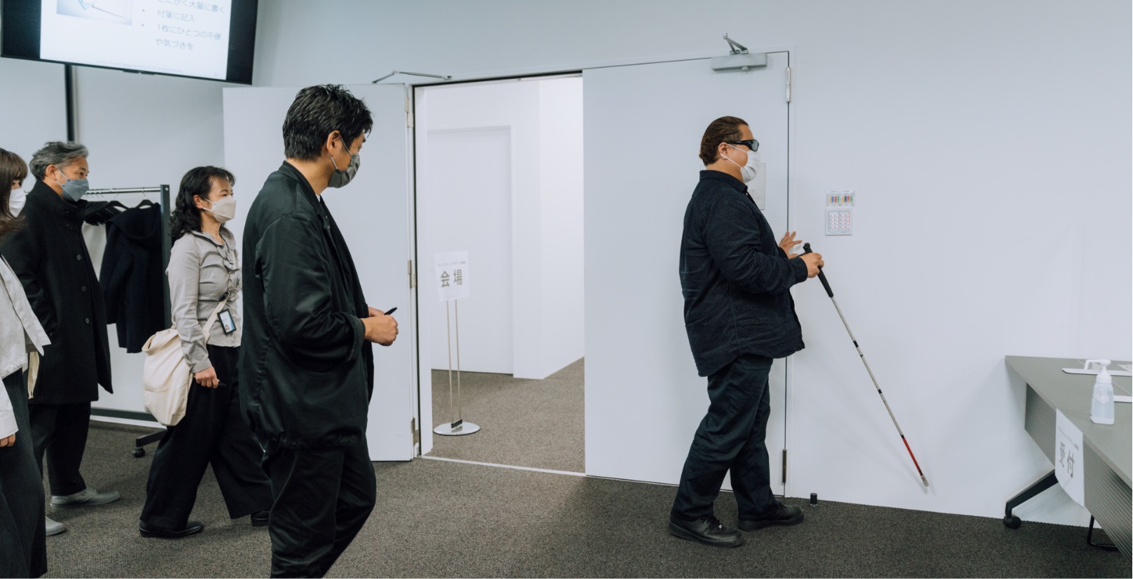 A group of people in a room, in which a blind person is touching the wall with his hand and white cane.