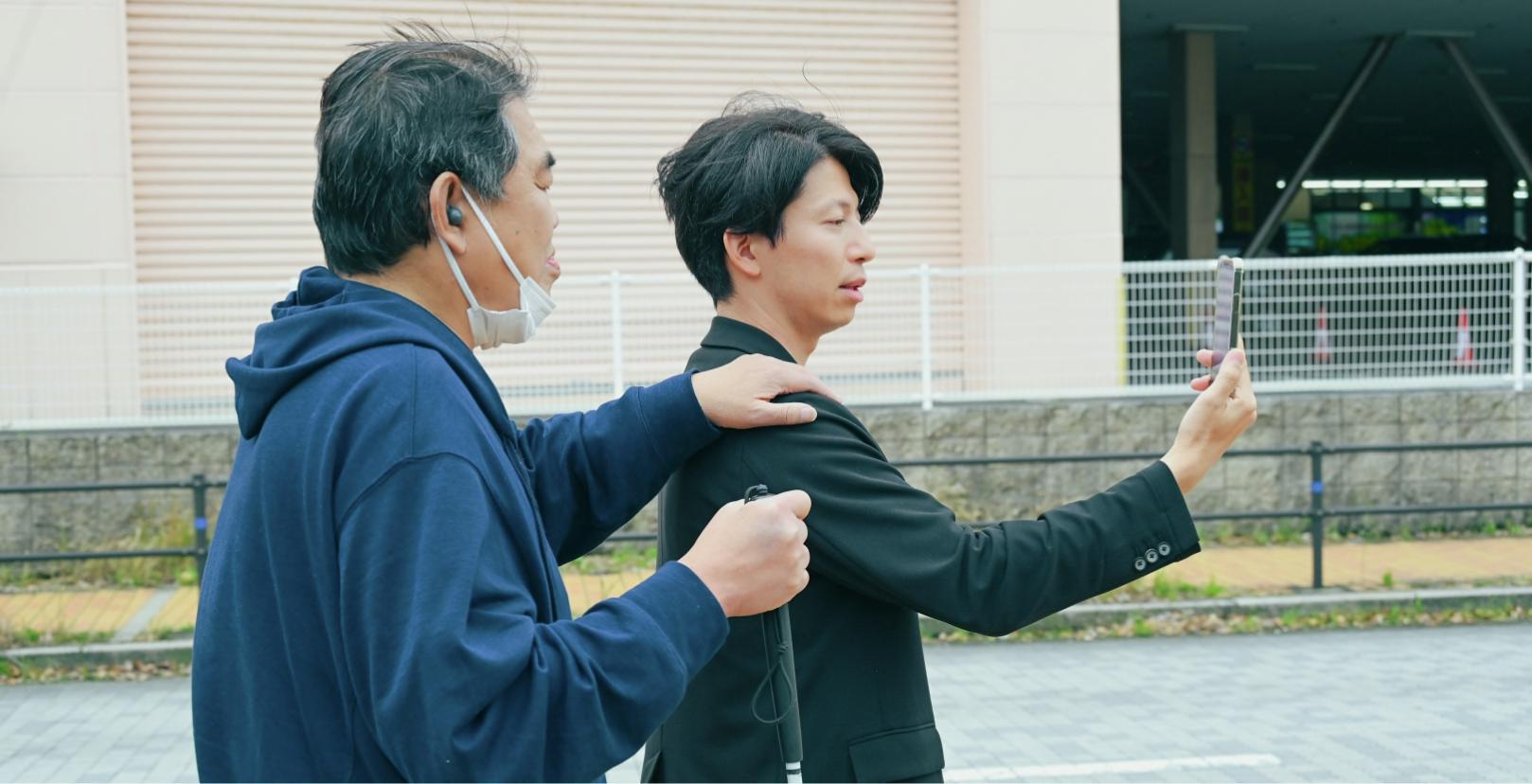 Shohei Takata is holding a smartphone, and another male with a white cane wearing LinkBuds is putting his left hand on Takata's shoulder